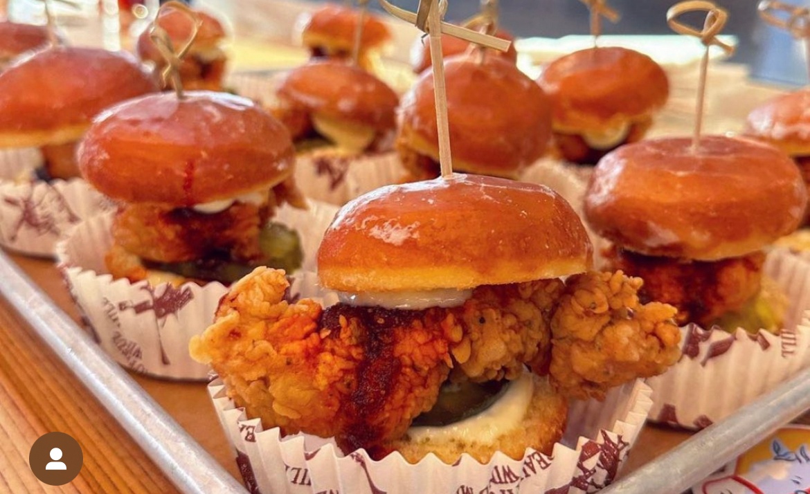 Mini fried chicken sliders in cupcake liners, arranged on a tray. Each slider has fried chicken, pickles, and a small bun, held together with wooden skewers.