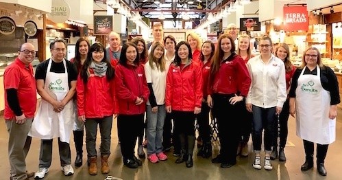 A group of people, some in red jackets and others in white aprons, pose together indoors at a market or food hall. They are standing in front of various food stalls with signs.
