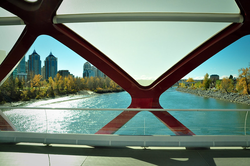 View through the red beams of a modern bridge, overlooking a river and city buildings with a clear sky and autumn trees lining the riverbank.