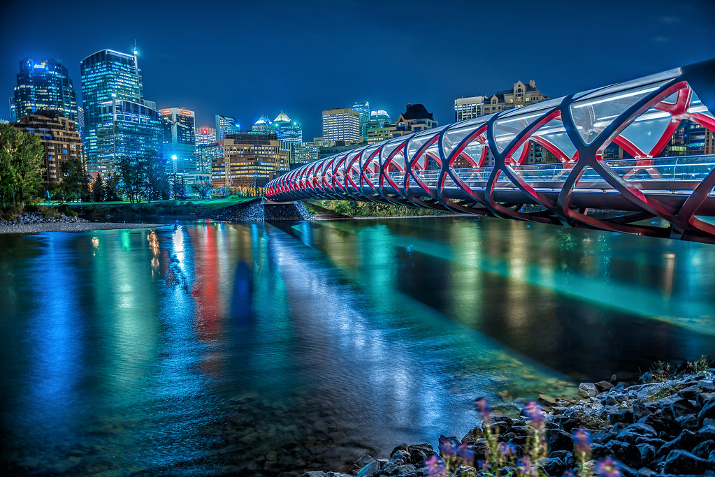 A modern, red-and-white truss pedestrian bridge is illuminated at night, reflecting on a calm river, with a city skyline in the background.