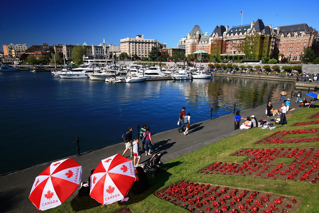 People walk along a waterfront path lined with boats and buildings. Red and white umbrellas with "Canada" printed on them and flower arrangements are visible in the foreground.