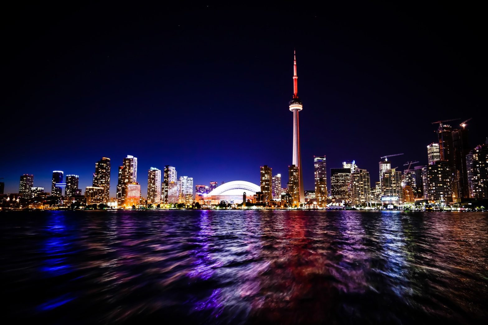 Toronto city skyline at night, featuring the CN Tower illuminated in red and white, and reflected lights on water.