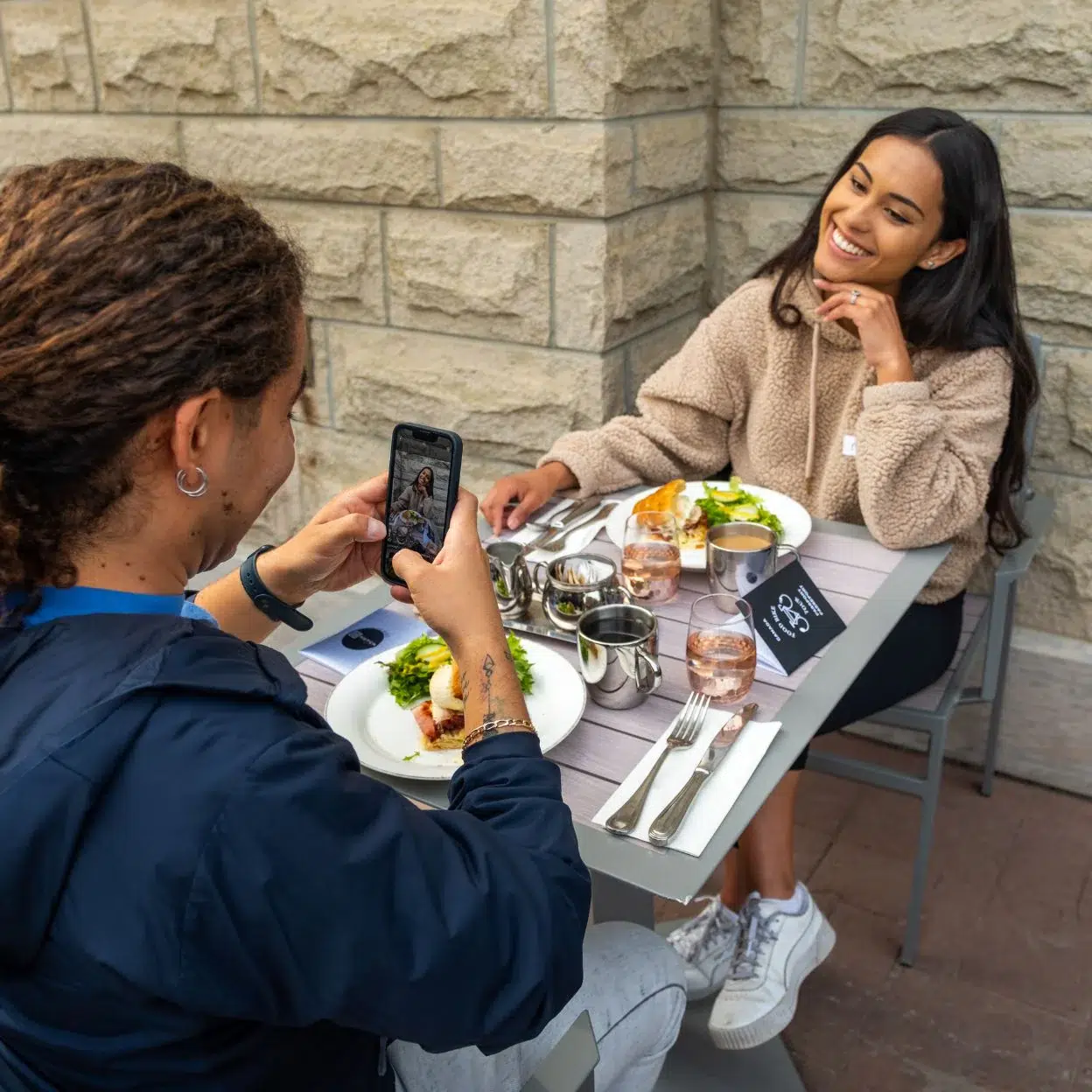 Two people seated at an outdoor dining table. One person takes a photo of the other, who is smiling. Plates with food and drinks are on the table.