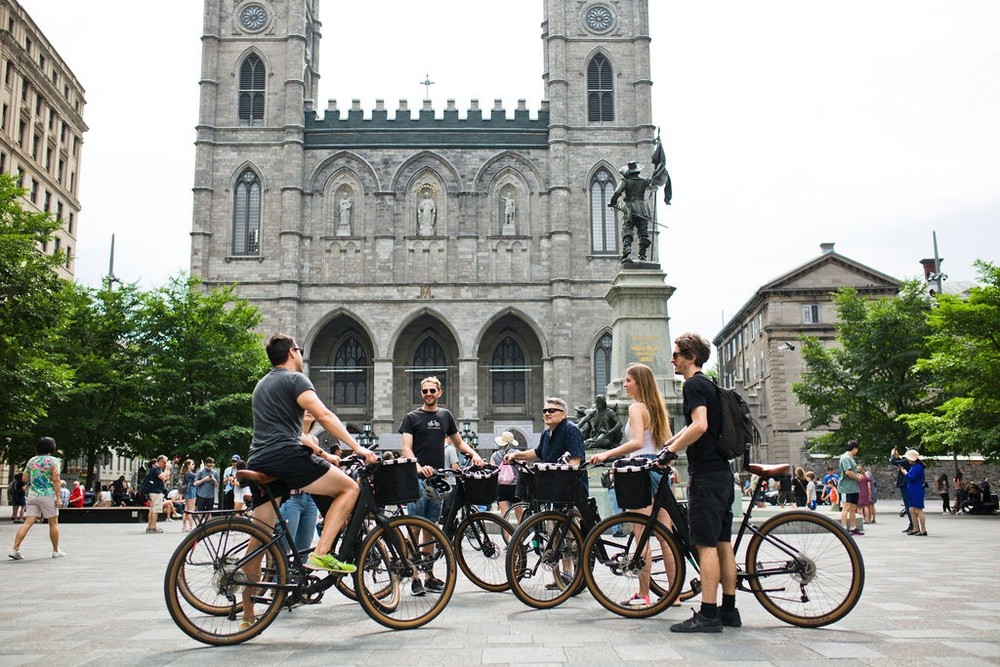 A group of people on bicycles are gathered in front of a large historic cathedral in a bustling plaza.