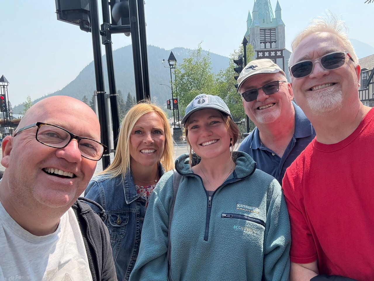 Five adults smiling and posing for a selfie outdoors in a mountain town with a clock tower and traffic lights in the background.