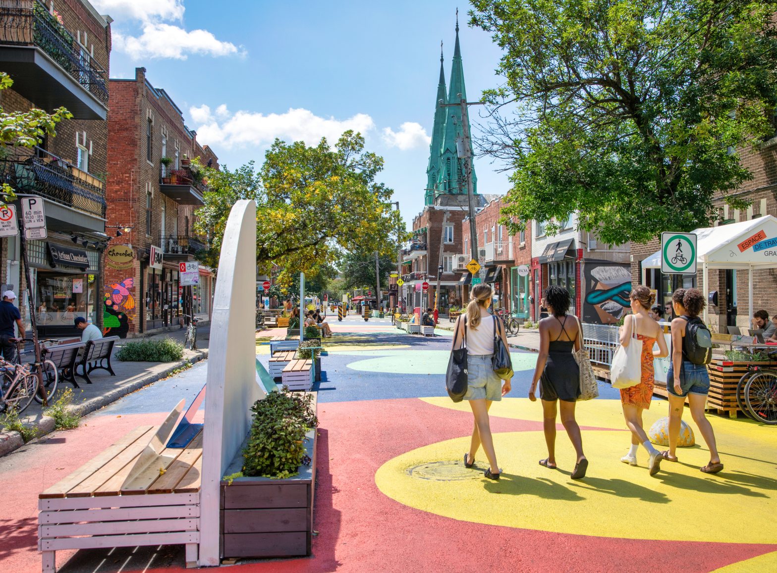 A group of four people walk along a colorful pedestrian street lined with shops, benches, trees, and a church with green spires in the background.