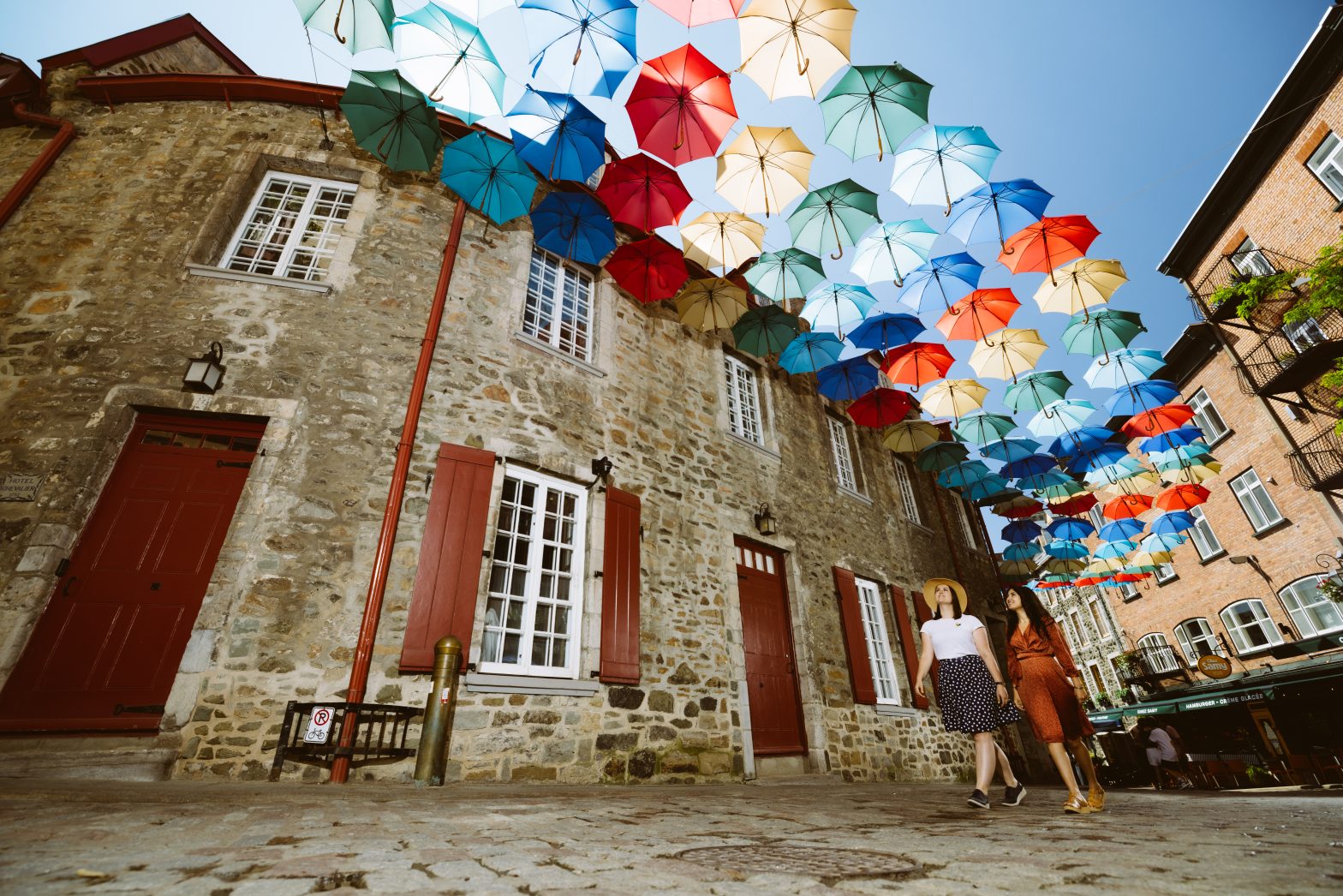 Two people walk on a cobblestone street under a canopy of colorful umbrellas hanging between historic stone buildings.