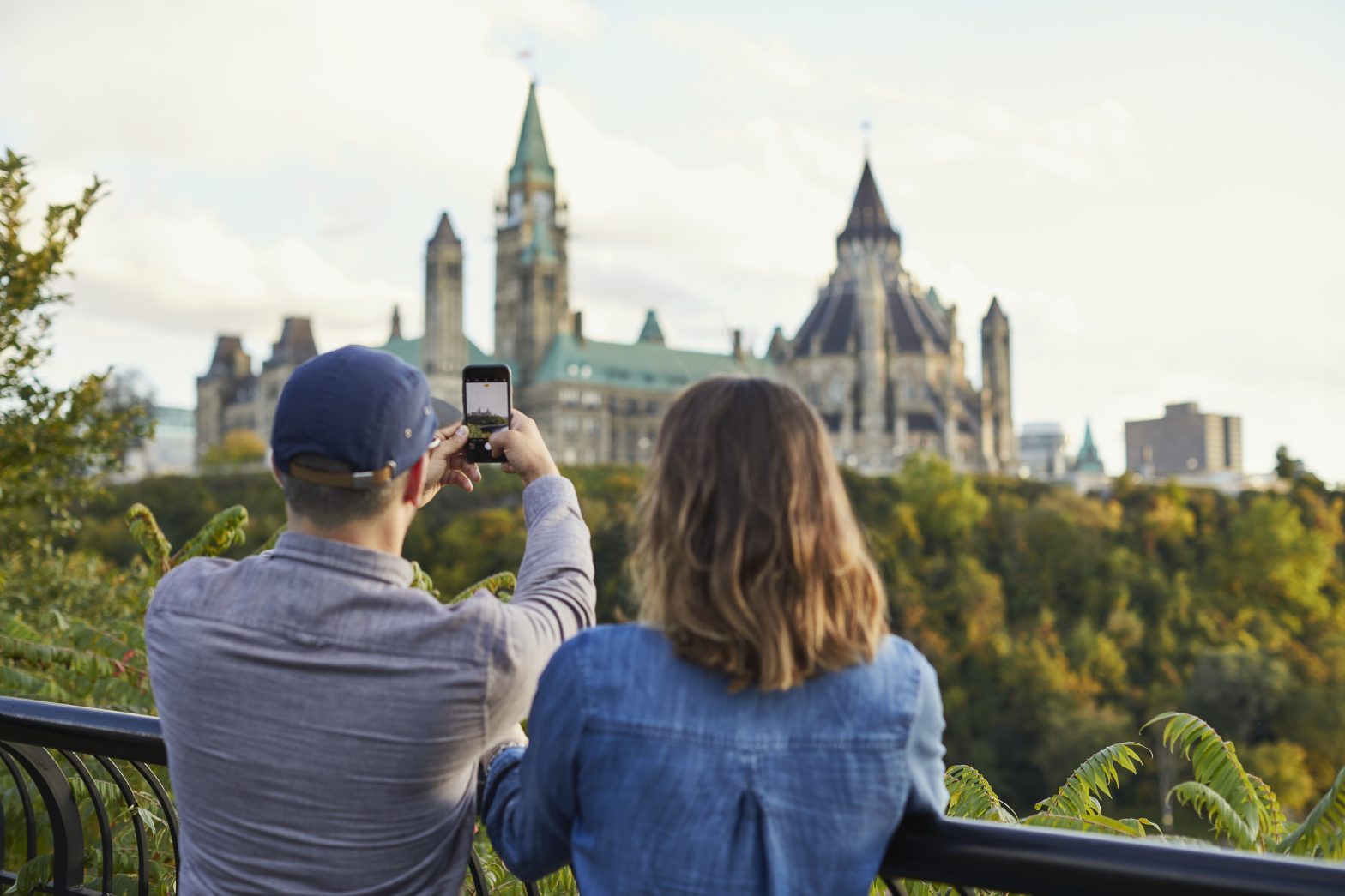 Two people stand on a railing, with one taking a photo of a large historic building surrounded by trees. The sky is partly cloudy.