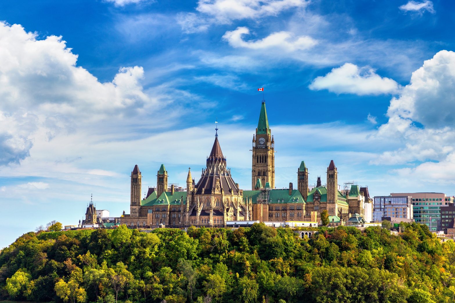 A distant view of Canada’s Parliament Buildings in Ottawa atop a tree-covered hill, under a partly cloudy blue sky.