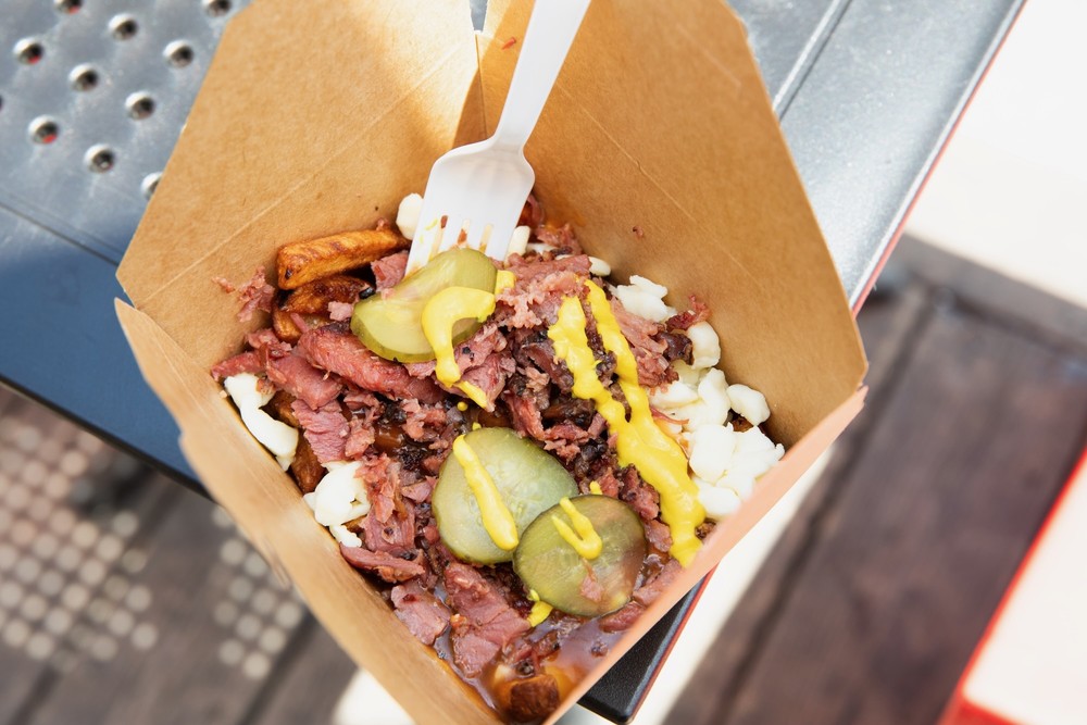 A takeout container filled with poutine, topped with shredded meat, pickle slices, and mustard. A white plastic fork is inserted into the meal.