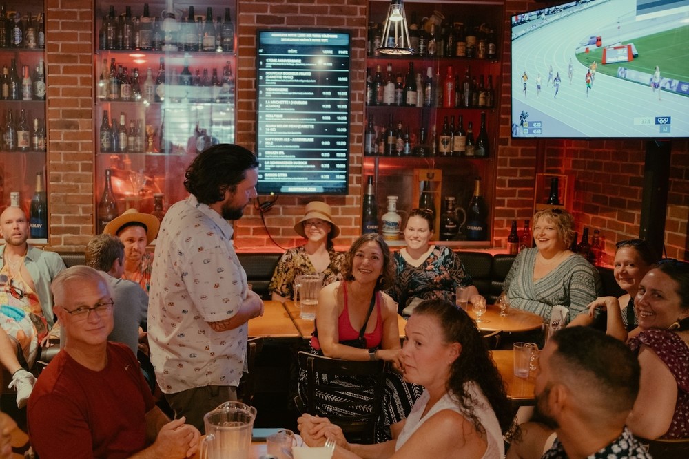 A group of people seated in a bar watching a sports event on a TV. The room has a brick wall, a large menu display, and shelves with bottles. A man stands and faces the seated group.