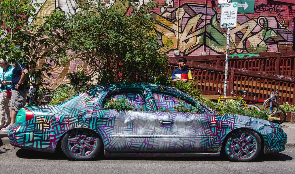 Car covered in colorful graffiti parked on the street, surrounded by plants and graffiti on nearby walls. People and a bicycle are visible in the background.