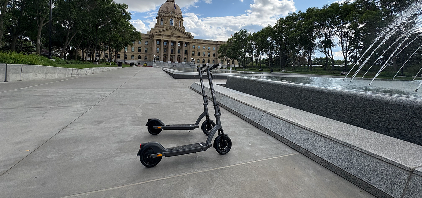 two electric scooters are parked on a wide sidewalk near a fountain, with a large government building and trees in the background.