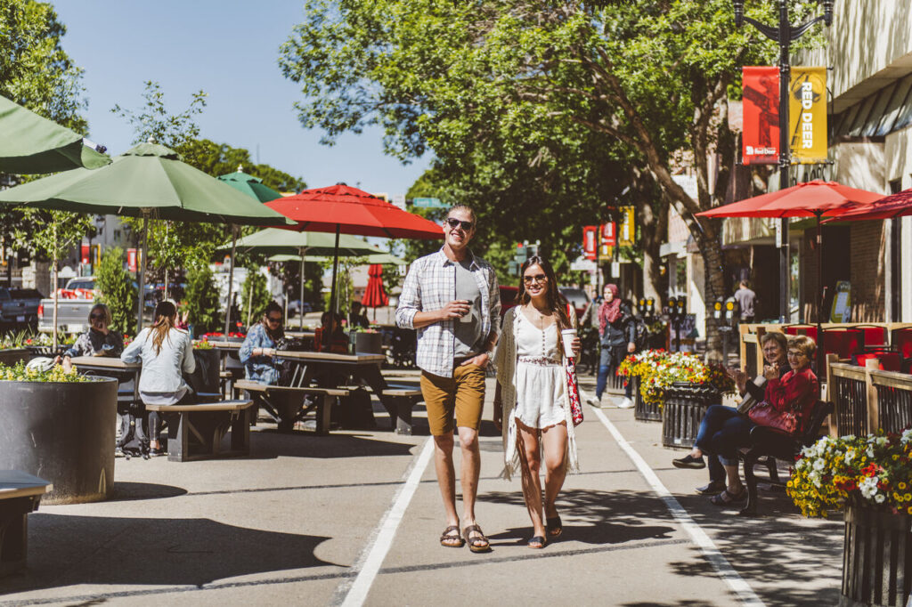 a man and woman walk down a pedestrian street lined with outdoor tables, umbrellas, and people dining on a sunny day.