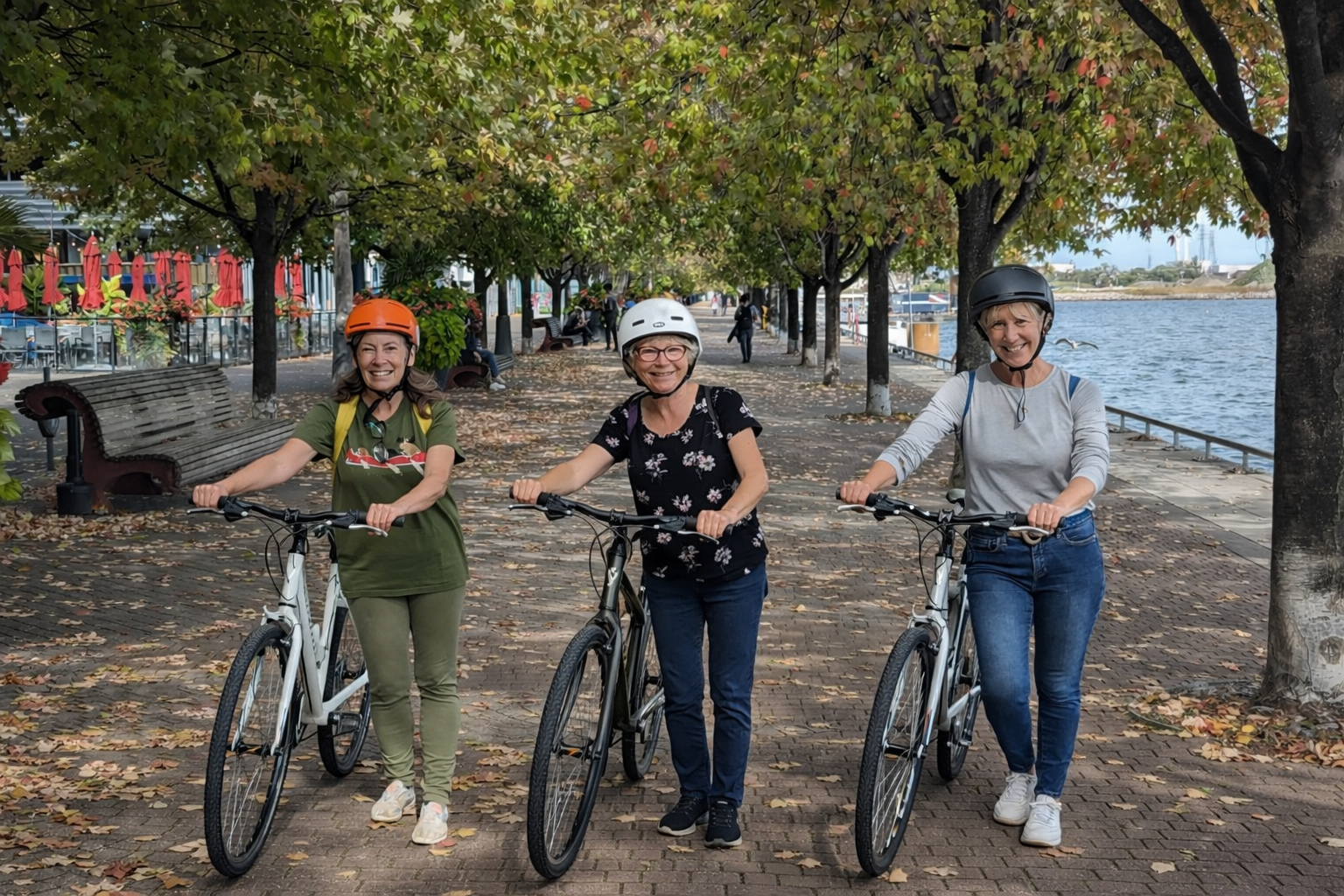 three women wearing helmets stand with bicycles on a tree lined path beside a body of water, smiling at the camera.