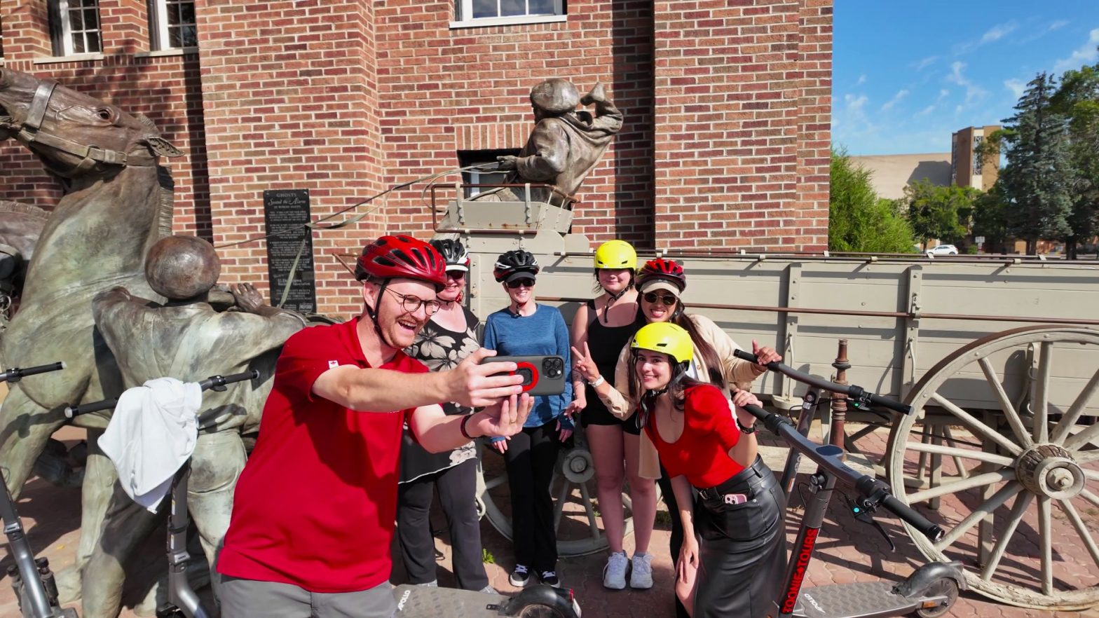 a group of six people wearing helmets take a selfie in front of a brick building and a bronze statue with scooters nearby.