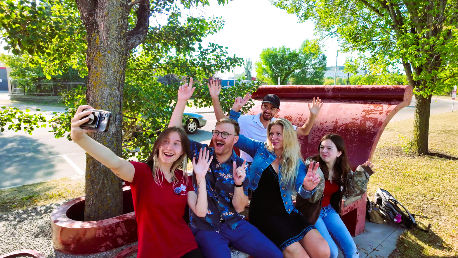 five people sit on a red bench outdoors, smiling and waving at a phone held up for a group selfie. trees and parked cars are visible in the background.