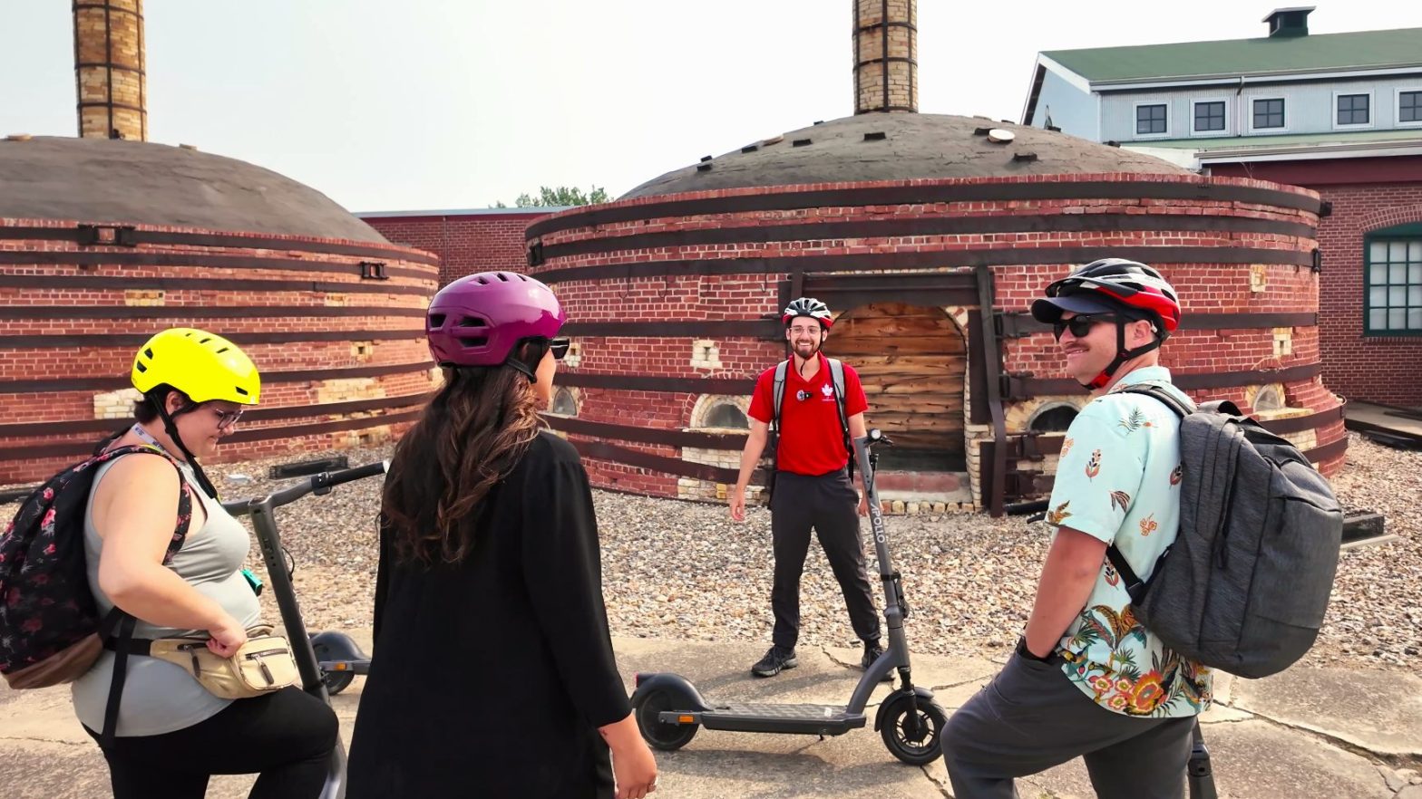 four people wearing helmets stand with electric scooters in front of round brick kilns, engaged in conversation on a sunny day.