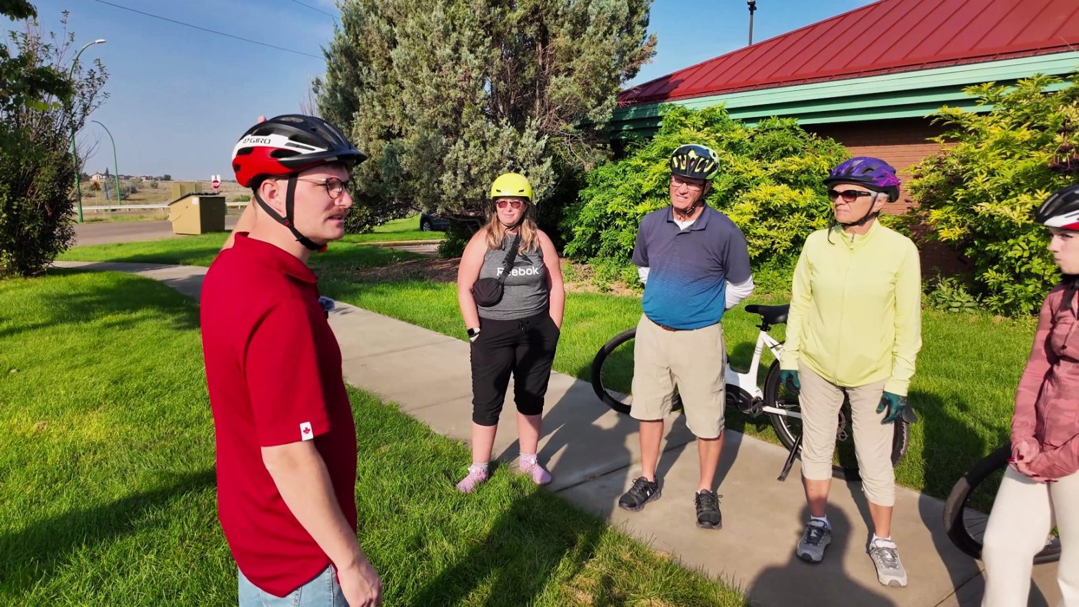 four people wearing bike helmets stand outside on grass near a sidewalk, talking. one bicycle is visible. there are trees and a building with a red roof in the background.