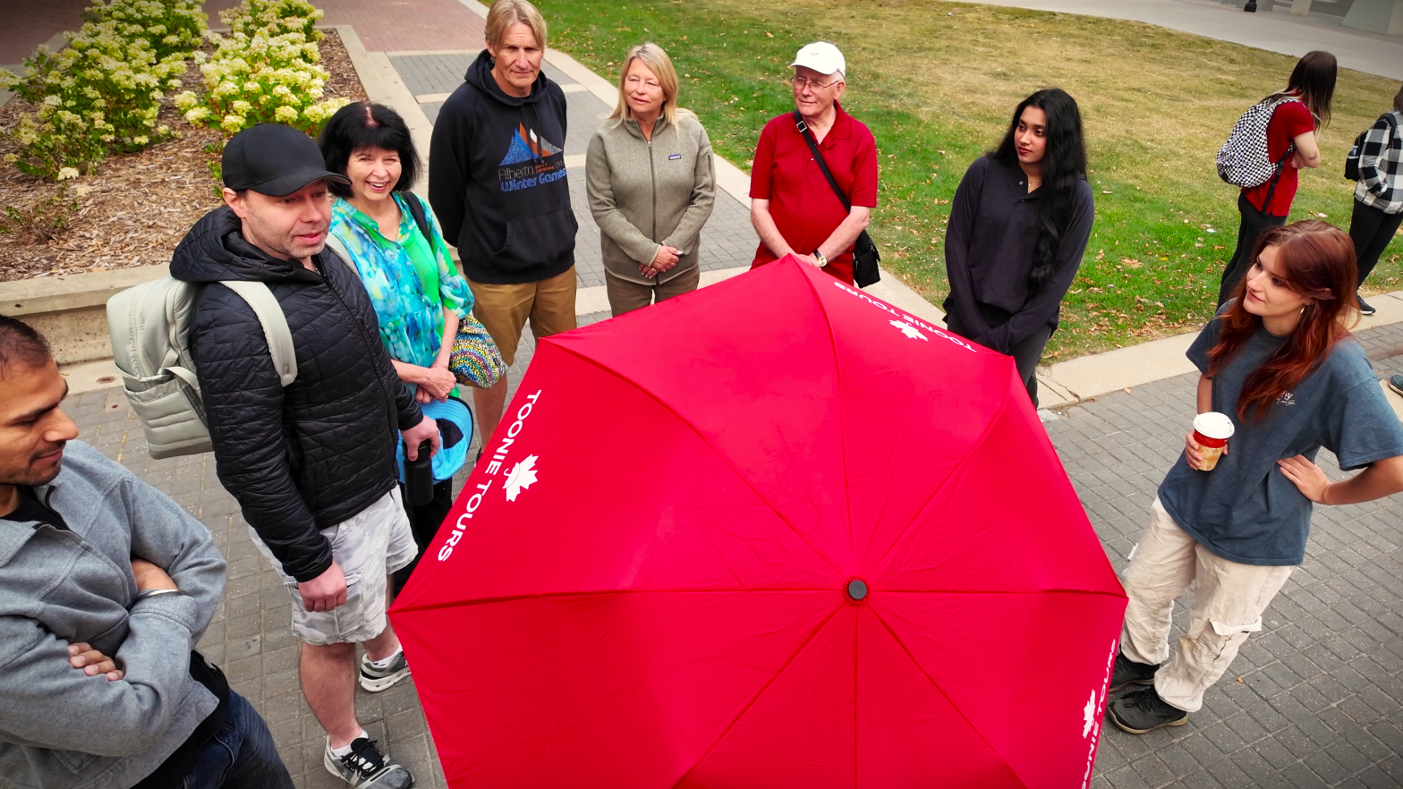 a group of eight people stand in a circle outdoors on a paved area, with a large red umbrella featuring maple leaf designs at the center.
